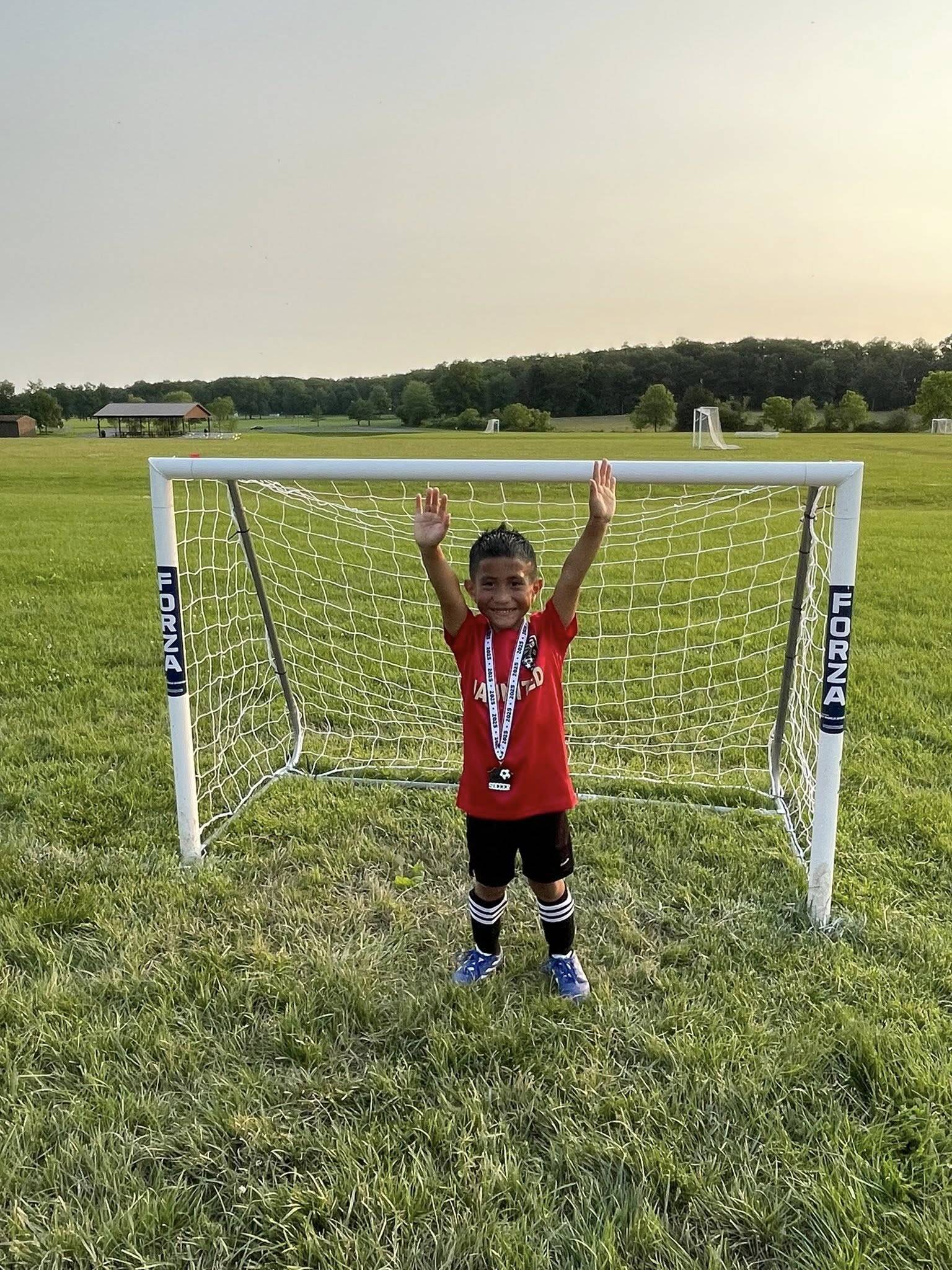 A young boy in a red shirt and blue sneakers stands in front of a soccer goal